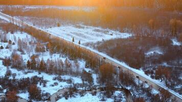 A straight road cuts through a snow covered landscape with sparse trees, a winding stream, and vehicles, illuminated by golden sunlight. video