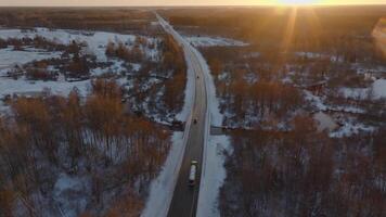 A straight road extends through a snowy terrain with leafless trees, open fields, and water bodies, illuminated by low golden sunlight on the horizon. video