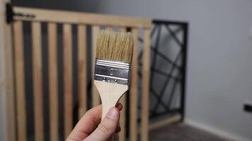 A hand holds a 2 inch natural bristle paintbrush. A wooden gate with vertical slats and a black geometric metal frame is visible in the indoor setting. video