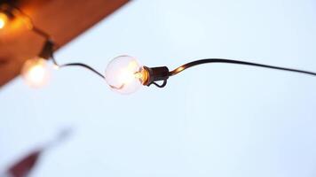 A glowing LED bulb on a black wire emits warm light with another bulb blurred in the background. A wooden structure and red object are partially visible. video