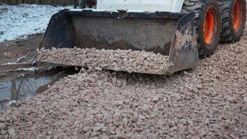 A small construction vehicle spreads crushed stones on a pathway in a snowy area, with a fenced section on the left and a wooden structure on the right. video