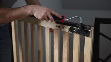 A person uses a small roller brush with a red handle to paint the top rail of a wooden slatted structure black, set against a neutral gray background. video