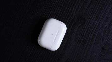 A white rectangular earbud charging case with rounded edges and an LED indicator rests on a dark textured wooden surface, emphasizing contrast. video