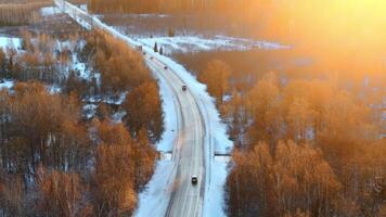 A winding road with a small bridge crosses a snow covered area bordered by golden hued leafless trees, with vehicles adding movement to the scene. video