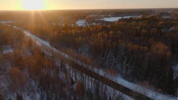 A snow covered road runs through a forest of deciduous and coniferous trees at sunset. A partially frozen body of water is visible in the distance. video