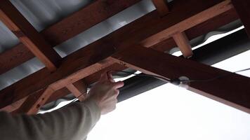 Close up of hands installing LED string light bulbs on a wooden terrace with triangular beams and a corrugated metal roof under an overcast sky video