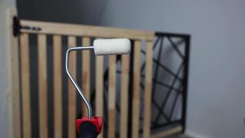 A paint roller with a red handle is in focus indoors, set against a blurred wooden safety gate with vertical slats and a black metal frame. video