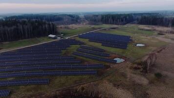 Rows of solar panels on a grassy field, a small building to the left, a pond to the right, a dirt road, and forested areas under an overcast sky. video