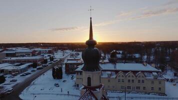A church with a tall spire topped by a cross stands in the foreground, surrounded by snow covered rooftops and streets, with a forested area in the background. video