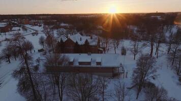 A snow covered scene during sunset featuring a brick building with a pitched roof, bare trees, pathways, and additional structures in the background. video