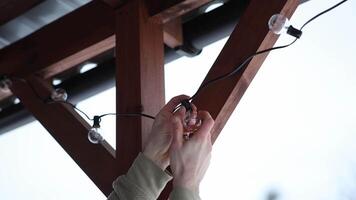 Two hands replace an LED bulb on a string of lights attached to a wooden terrace structure, with wooden beams and a tiled roof in view. video