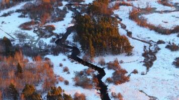 A winding river flows through a snow covered landscape, bordered by sunlit trees and open fields. Evergreen clusters contrast with the white snow. video