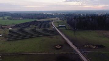 A large solar panel array spans open fields divided by a dirt road, bordered by grassland, dense forest, and a distant body of water under cloudy skies. video