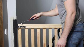 An individual uses a tool to secure a wooden safety gate with black metal hinges at the top of a staircase. A light switch is visible on the wall. video