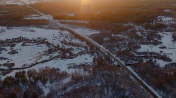 A long road traverses a snowy landscape with sparse trees and winding streams, illuminated by the golden light of a setting sun. video