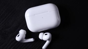 Pair of white wireless earbuds with matching charging case on a dark surface. One earbud lies on its side, while the other stands upright. video