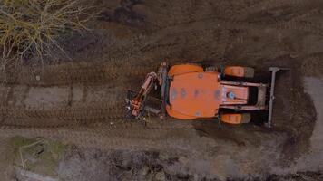 An orange excavator operates on a construction site, digging into dark soil. Surrounding areas show patches of grass and dirt contrasting the work zone. video