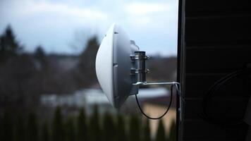 White dome shaped WiFi router mounted on house wall with metal brackets, set against evergreen trees, residential structures, and an overcast sky. video
