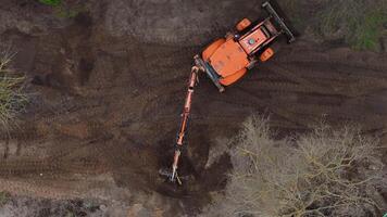 An orange excavator with an extended arm on a dirt surface, surrounded by sparse vegetation, including leafless trees and evergreen branches. video