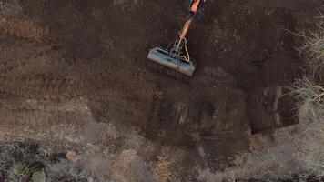 An orange excavator operates on dark soil, surrounded by tire tracks. Sparse vegetation and bare branches frame the edges of the construction site. video
