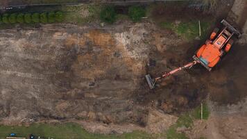 An aerial view shows an orange excavator working on a construction site, bordered by grass and sparse vegetation, with visible fencing and materials. video