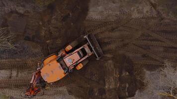 An aerial view shows an orange tractor on a dirt covered site with visible tire tracks, surrounded by sparse trees with bare branches in a natural setting. video