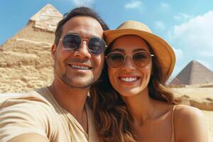 Smiling couple taking a selfie at the Pyramids, wearing sunglasses and hats, exuding joy and adventure under a bright blue sky. photo