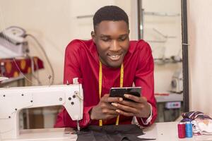 African tailor using his phone while working in his workshop. dressed in modern attire checking orders online photo