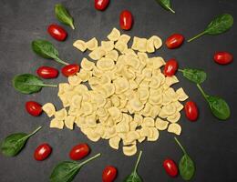 Uncooked tortellini pasta with fresh cherry tomatoes and spinach leaves on a dark, textured surface. Flat lay shot of ingredients arranged in a scattered pattern photo