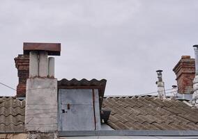 A close-up shot of several weathered chimneys on a rooftop, vary in design and material, some made of brick, others of concrete or metal, showcasing signs of age and exposure to the elements. photo