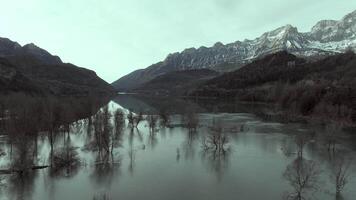 Aerial shot - Mountain landscape revealing pristine water surface mirroring snow capped peaks, leafless trees, and surrounding wilderness of Ibon de los Banos in winter season video