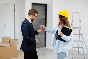 Designer in helmet with documents giving keys from apartment to young businessman while standing in room photo