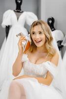 Bride-to-be wearing a wedding dress at a fitting room in a salon, choosing shoes for a wedding and having fun using a shoe as a phone. photo