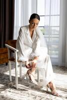 Bride wearing white suit and veil tying elegant high heels sitting on chair in hotel room. photo