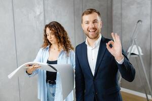 A young designer discusses the plans and design of the room with the customer. The customer is engrossed in work and shows the ok gesture. The concept of repair on a modern design photo