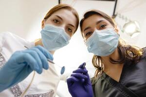 From below female dentists with mirror and device bending over patient during appointment in clinic. photo