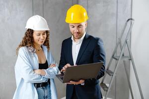 Young woman contractor is talking to customer in helmet, and demonstrates sequence of repairs in living room on laptop. Meeting of landman of apartment with engineer or architect. photo