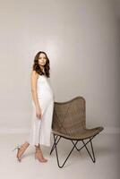 Pregnant Model Posing in White Dress with Wicker Chair in Studio photo