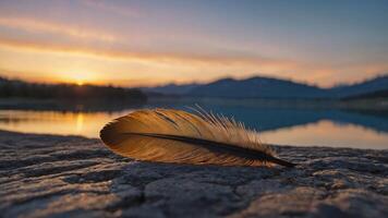 A feather rests on a rocky surface with a serene sunset and mountains reflected in the water. photo