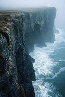 Dramatic coastal cliffs meet turbulent ocean waves in misty weather photo