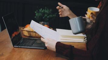 A woman is holding a cup of coffee while sitting at a table with a laptop video