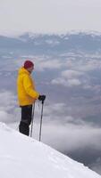 A person dressed in a bright yellow jacket stands on a snowy slope, gazing at the expansive landscape covered in clouds and mountains in the distance. Vertical footage video