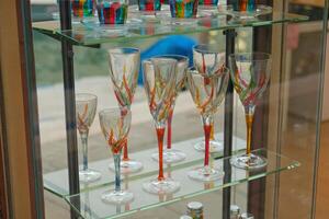 Elegant Murano glassware in Venetian style displayed on glass shelves in showcase store. Handcrafted artistic wine glasses and tumblers with vibrant multicolored designs, reflecting light. photo
