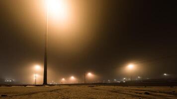 Streetlights cast an eerie glow through dense fog on a quiet, empty road at night, creating a moody and cinematic urban atmosphere. photo