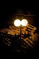 A glowing vintage street lamp stands against a dark backdrop, casting light over scattered wooden pallets. The eerie contrast between warmth and decay creates a mysterious, cinematic atmosphere. photo