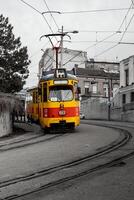 A classic yellow and red tram, marked with route number 11, makes a gentle turn along curved tracks in an urban setting. The muted tones of the cityscape contrast with the tram's vibrant colors. photo