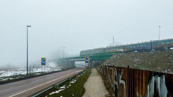 A colorful graffiti-covered train moves across a misty green bridge over a quiet road, surrounded by a foggy winter landscape. photo
