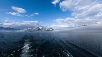 A breathtaking view from a boat, showcasing deep blue Arctic waters, mist-covered snow-capped mountains, and a vast sky with scattered clouds. photo