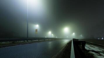 A desolate highway shrouded in thick fog, with glowing streetlights casting an eerie atmosphere. The misty road disappears into the unknown, evoking mystery and solitude. photo