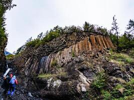 A group of hikers admires impressive hexagonal basalt columns in a remote canyon, showcasing unique geological formations surrounded by lush greenery and a small waterfall photo
